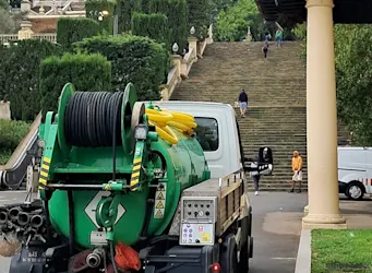 Camión verde de mantenimiento equipado con mangueras estacionado al pie de unas escaleras en un entorno verde y arquitectónico, posiblemente en un parque o sitio histórico.