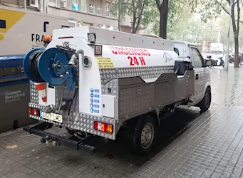 Camión de servicios especializados en desatascos 24 horas estacionado en un entorno urbano con vegetación y edificios al fondo.